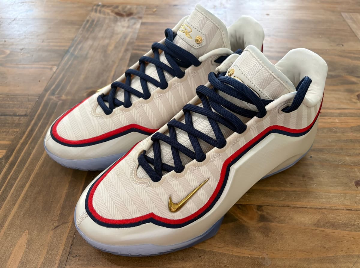 Nike LeBron 23 Old Glory with a cream herringbone upper, red and navy piping, navy laces, a gold Swoosh and an icy blue outsole, on a wood floor